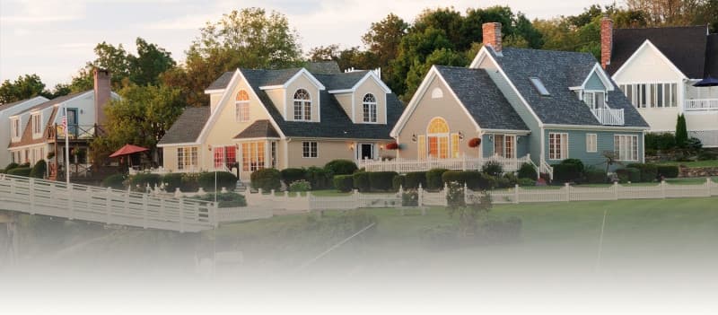 A row of several houses with varying architectural styles is visible along a waterfront. Green lawns and white fences are present in the foreground, with trees in the background.